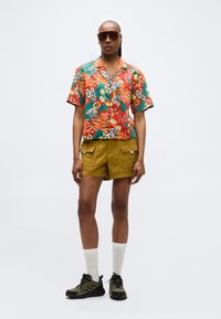 Γυναίκα με μεγάλα καφέ γυαλιά ηλίου, κοντομάνικο πουκάμισο με τροπικό σχέδιο, μουσταρδί cargo σορτς, άσπρες κάλτσες και αθλητικά παπούτσια σε ελιά πράσινο χρώμα.