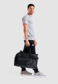 Bolsa de deporte negra con un gran logotipo, hecha de material durable, con asas resistentes y un bolsillo lateral de malla, llevada por un hombre en ropa deportiva.