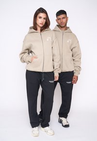 Beige zip-up hættetrøjer med lommer, kombineret med sorte joggere med hvide logo detaljer. Glat stof, afslappet pasform, casual design.