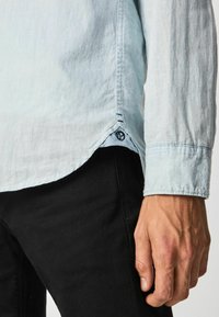 Camicia a maniche lunghe blu chiaro con orlo curvo, caratterizzata da cuciture scure e un piccolo logo sul lato. Pantaloni neri parzialmente visibili.