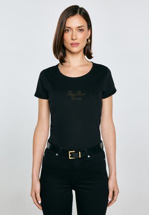 Žena s hnědými vlasy po ramena, oblečená v přiléhavém černém tričku se zlatým logem a černých kalhotách s černým páskem, stojící na jednobarevném pozadí.