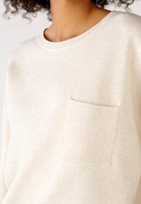 Osoba v světle béžovém pleteném svetru s kulatým výstřihem a malou náprsní kapsou, zobrazená od pasu nahoru s viditelným krkem a horní částí trupu.