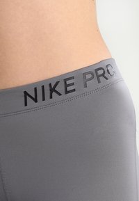 Zbliżenie na talię osoby noszącej szare legginsy sportowe Nike Pro z czarnym logo na pasie, na jednolitym tle.