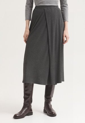 Personne portant une jupe midi gris foncé avec un pli à l'avant et de grandes bottes en cuir marron foncé, debout devant un fond clair uni.