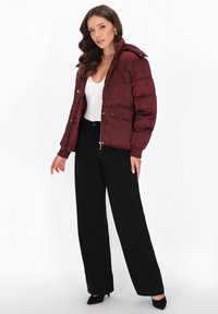 Giacca a vento bordeaux con zip frontale, due tasche laterali e cappuccio rimovibile. Indossata sopra un top bianco e abbinata a pantaloni neri a gamba larga.