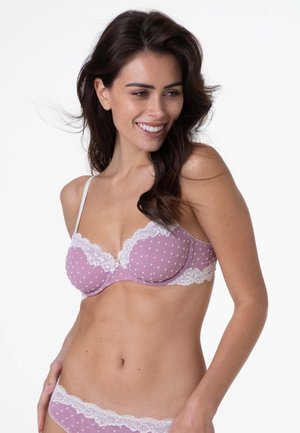 Donna che sorride, indossando un set di lingerie a pois rosa con rifiniture in pizzo bianco, posando su uno sfondo bianco uniforme.