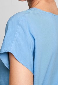 Top bleu clair en tissu texturé et respirant, doté de manches courtes et d'un col arrondi, avec des détails de couture au niveau des coutures.