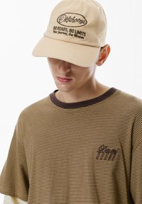 Cappellino da baseball beige con testo ricamato marrone, abbinato a una maglietta a righe marroni e gialle con un piccolo badge logo.