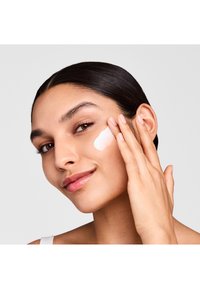 Feuchtigkeitspflege auf die Wange aufgetragen; eine glatte, weiße Creme mit einem glänzenden Finish. Das Model hat glattes, dunkles Haar und einen natürlichen Teint.