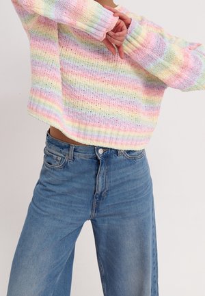 Person iført pastelregnbue-stribet strikket sweater og højtaljede blå jeans med armene krydset foran torsoen.