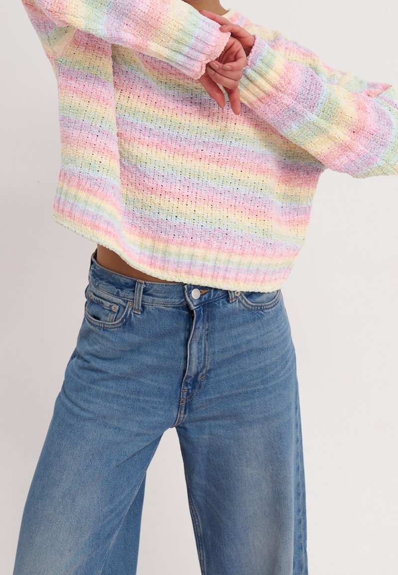 Personne portant un pull en tricot rayé arc-en-ciel pastel et un jean bleu taille haute, bras croisés devant le torse.