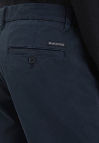 Donker marineblauwe broek met een gladde textuur, een achterzak met een knoop en een zwart label met de tekst "Marc O'Polo."