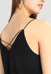 Svart top med tunna axelband och korsettsydd ryggdesign. Tyget ser slätt och lätt ut med en subtil glans.