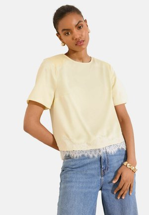 Kobieta ubrana w kremową bluzkę z krótkim rękawem i koronkową lamówką oraz niebieskie dżinsy, dopełniona złotymi kolczykami kołami i złotą bransoletką.