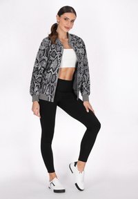 Fekete-fehér kígyóbőr mintázatú bomber dzseki, bordás mandzsettákkal és elöl cipzárral, fekete leggings-szel és fehér sportcipőkkel párosítva.