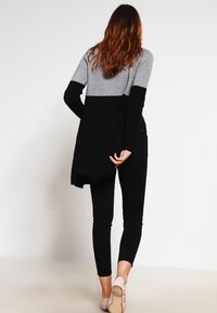 Zwarte en grijze lange-sleeve cardigan met een gespleten achterkant ontwerp, gedragen over zwarte skinny jeans en lichtgekleurde schoenen.
