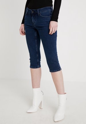Kvinde iført mørkeblå cropped skinny jeans med en sort langærmet top og hvide hælede ankelstøvler, stående mod en ensfarvet baggrund.