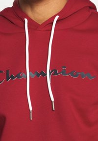 Sweat à capuche rouge en mélange de coton, avec un logo "Champion" argenté brillant et des cordons blancs à embouts métalliques. Texture lisse.