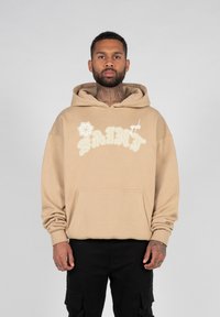 Hoodie beige en coton, avec une poche frontale et un design brodé "SAINT" en blanc. Comprend une capuche à cordon et des manches longues.