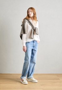Beige Langarmoberteil mit einem gerippten grauen Pullover über den Schultern, hellblaue lässige Jeans und cremefarbene Sneaker mit rosa Akzenten.