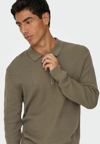 Olive roheline kootud pullover, millel on lukk-kaelus, pikal varrukatel ja tekstuurilisel kootud kujundusel. Materjal näeb välja pehme ja kerge.