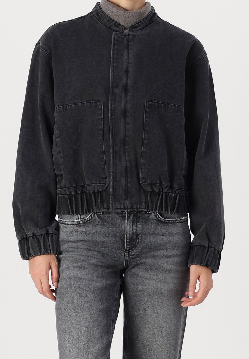 Personne portant une veste courte noire avec de grandes poches avant et des poignets élastiques, associée à un jean gris délavé et un col roulé gris.