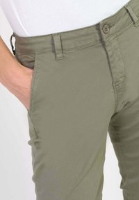 Close-up of a person wearing olive green pants with a white top, hand casually placed in the front pocket on the left side.