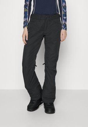 Osoba ubrana w czarne spodnie narciarskie i czarne zimowe buty stojąca na białym tle.