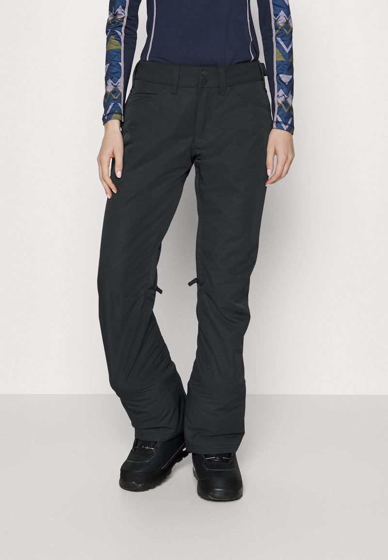 Personne portant un pantalon de ski noir et des bottes d'hiver noires debout sur un fond blanc.
