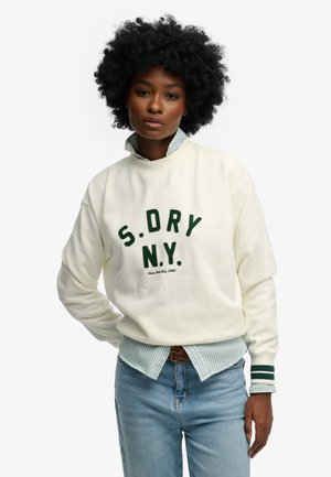 Ung kvinde med krøllet hår iført en hvid sweatshirt med teksten "S.DRY N.Y.", stribet skjorte under og lyseblå jeans, står stille.