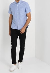 Camicia celeste chiaro a maniche corte con bottoni e taschino sul petto, abbinata a pantaloni neri attillati e sneakers bianche. Design semplice, materiale in cotone.