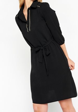 Femme en robe noire longueur genou avec manches trois-quarts, col, ceinture nouée devant et surpiqûres blanches au col et à la patte de boutonnage.