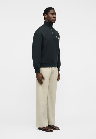 Άνδρας που στέκεται πλαγίως φορώντας μαύρο πουλόβερ με φερμουάρ στο ένα τέταρτο, μπεζ παντελόνια με φαρδιά μπατζάκια και σκούρα καφέ σανδάλια σε απλό φόντο.