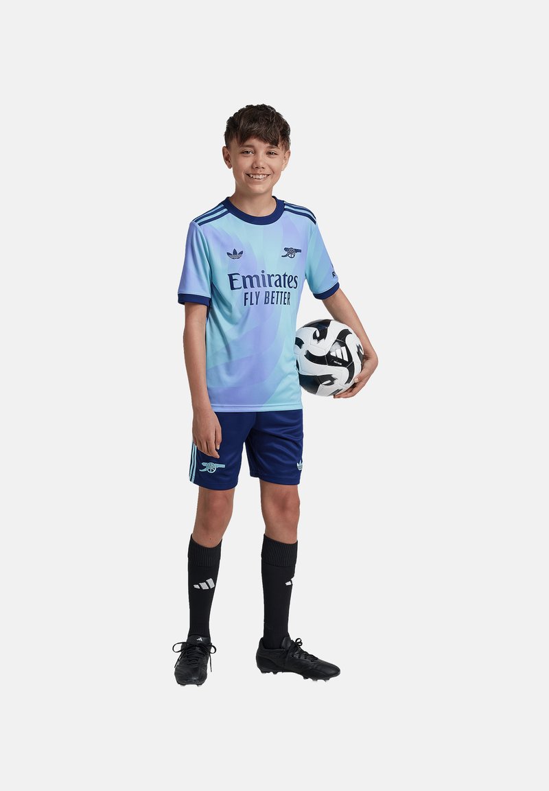 Valk blue soccer jersey dengan kerah navy, teks "Emirates Fly Better", dipasangkan dengan celana pendek navy, kaus kaki hitam, dan sepatu kets hitam; memegang bola sepak bermotif.