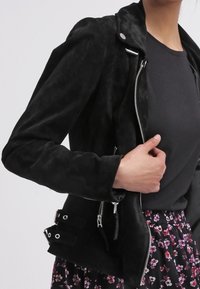 Veste biker en daim noir avec une silhouette ajustée, des fermetures éclair argentées, des boutons-pression et des sangles de taille réglables, portée sur un haut gris.