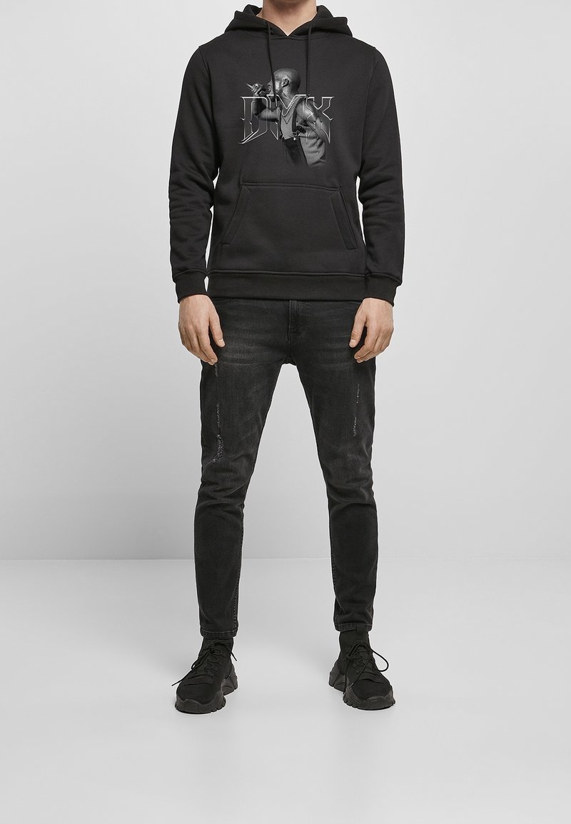 Homme portant un sweat à capuche noir avec un motif de chanteur, un jean noir et des baskets noires, debout contre un mur et un sol gris.