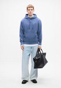 Giovane uomo con felpa blu e jeans larghi chiari, che tiene in mano una grande borsa di pelle nera, in piedi su uno sfondo bianco uniforme.