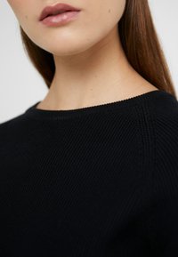 Pull en tricot noir avec une finition texturée, comportant un col rond simple et des détails en fins côtes le long du col.
