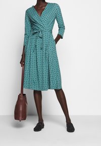 Robe portefeuille turquoise avec un motif tourbillonnant, manches trois-quarts et taille nouée. Le modèle l'associe à un sac en cuir marron et des chaussures noires.