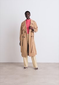 Beige trenchcoat met een dubbel-breasted ontwerp, grote kraag en zijzakken. Gedragen over een roze top en beige broek, met een gladde textuur.