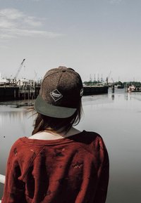 Brown cap with a green brim featuring a diamond logo and side pin. Person wearing a maroon sweater, standing by a body of water with cranes.