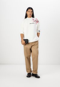 Sweatshirt blanc avec imprimé floral et logo sur la poitrine, associé à un pantalon large beige et des chaussures noires. Design simple, coupe décontractée.