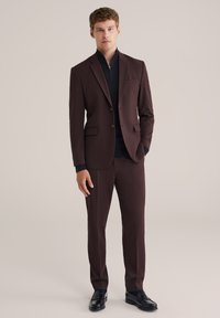Hombre de pie vistiendo un traje marrón oscuro con blazer de dos botones, suéter negro con cremallera y mocasines negros, contra un fondo beige.