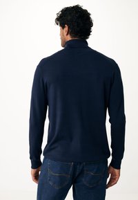 Marineblauer Pullover mit langen Ärmeln, hohem Kragen und gerippten Bündchen. Aus einem glatten Strickstoff gefertigt und kombiniert mit dunklen Jeans mit Steppdetails.