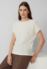 Witte textuur short-sleeve shirt met een golvend patroon, ronde halslijn en zachte stof. Gecombineerd met een bruine broek op een lichte achtergrond.