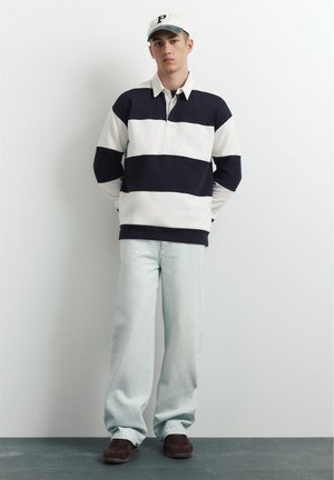 Jeune homme portant un t-shirt à manches longues blanc et noir rayé, un jean bleu clair, des chaussures marron et une casquette blanche avec la lettre P noire.