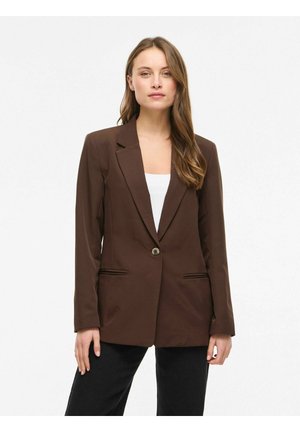 Kvinne med langt hår iført en brun blazer med én knapp over en hvit topp og svarte bukser, stående mot en ensfarget lys bakgrunn.