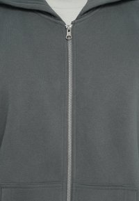 Felpa zip grigia realizzata in tessuto morbido, con zip frontale, grande cappuccio e tasche a marsupio, dal tocco liscio.