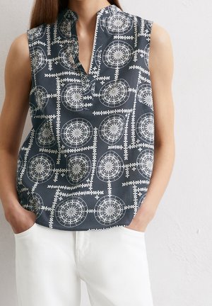 Haut sans manches bleu marine avec des motifs géométriques et floraux circulaires blancs, porté avec un pantalon blanc, mains dans les poches, sur un fond neutre.
