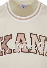 Cremefarvet mesh t-shirt med ribbet rund hals. Har "KANI" i fed brun og lys pink skrifttype, samt "originals" i hvid kursiv.
